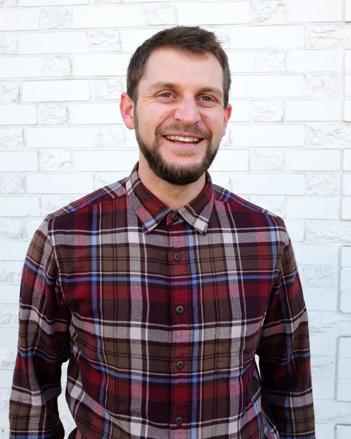 Photographic portrait of Patrick Hayden smiling, wearing a maroon and blue flannel shirt and standing in front of a white brick wall. Patrick has short brown hair and a beard.